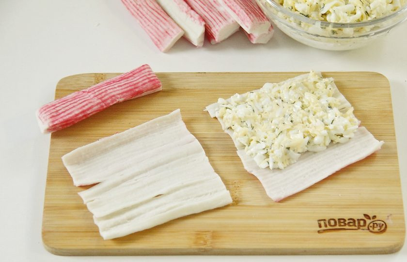 1. Этап. Новогодние рулетики из крабовых палочек: Крабовые палочки, если необходимо разморозьте и аккуратно разверните, стараясь не порвать. Если крабовые палочки плохо разворачиваются, то отправьте их буквально на 5-10 секунд в микроволновку, после чего мягко помните руками. Положите на развернутую палочку немного начинки, отступив от края около 1,5-2 см.