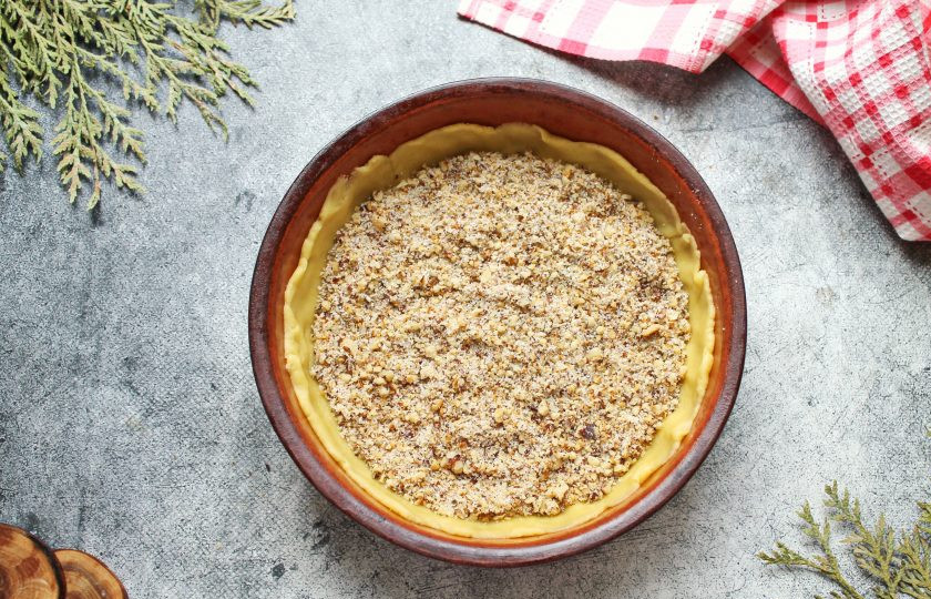 1. Этап. Ботлихский пирог: Первую часть теста раскатайте в круг и выложите в форму диаметром 18 сантиметров. Распределите тесто по дну и бокам формы. Всыпьте ореховую начинку, отложив две столовые ложки.