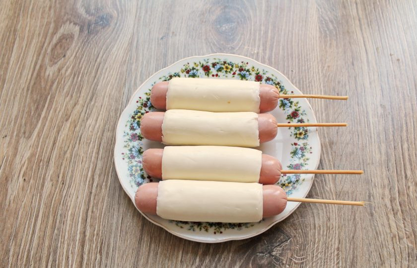 1. Этап. Сосиски в панировке с сыром: Воткните деревянную шпажку практически до конца. Выложите в тарелку сырным швом вниз и отправьте в холодильник на 20 минут.