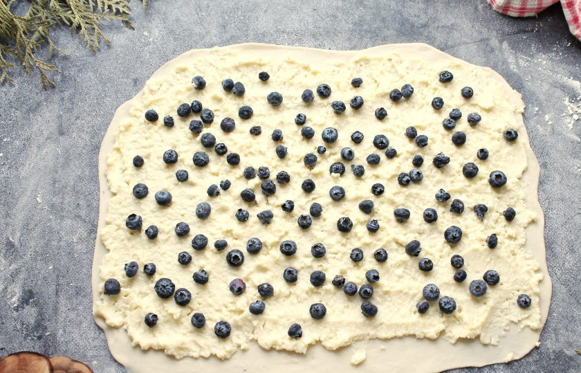 1. Этап. Булочки с голубикой: Рабочую поверхность посыпьте немного мукой и раскатайте тесто в прямоугольник толщиной 0.5-0.8 сантиметра и смажьте творожной массой, оставив один край пустым. Посыпьте творожный слой ягодами голубики.
