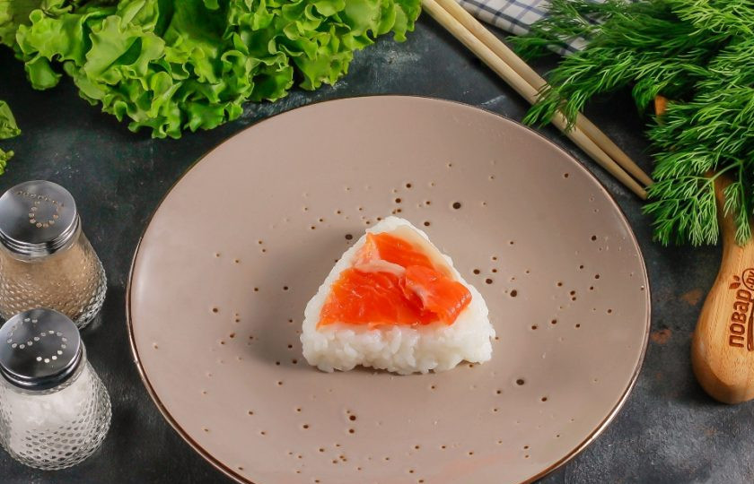 1. Этап. Онигири с лососем: Из теплого риса вылепите треугольник и выложите на него нарезку лосося. Залепите сверху рисом, придавая форму треугольника с округлыми углами.