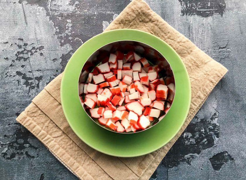 1. Этап. Салат Безумный с крабовыми палочками: Возьмите сервировочное кольцо, выложите его на тарелку. Крабовые палочки мелко нарежьте и выложите нижний слой.