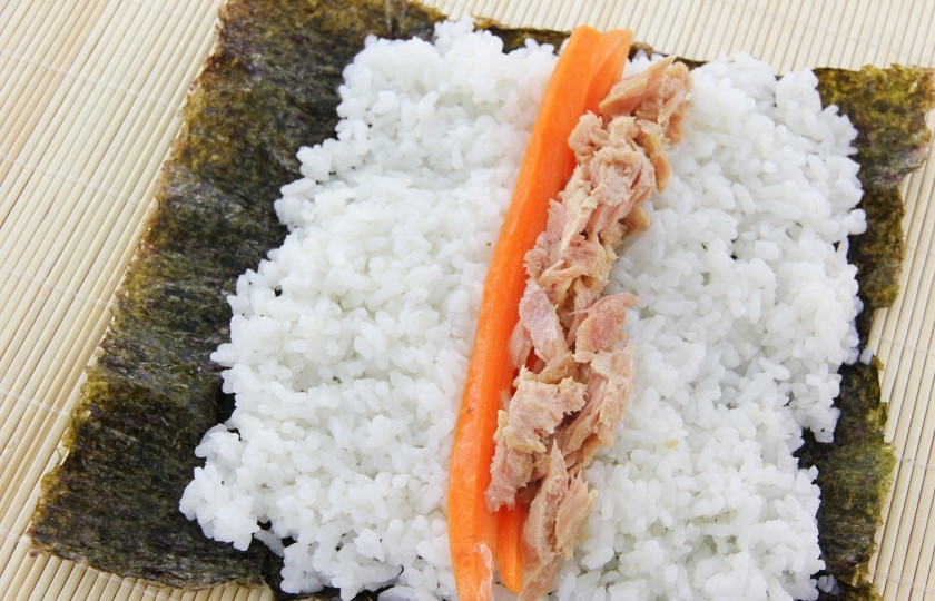 1. Этап. Роллы с консервированным тунцом: Выложите на лист нори рис. С левой стороны должно остаться немного пустого места, чтобы было удобно закрепить лист при сворачивании ролла. Сверху уложите морковь и тунец.