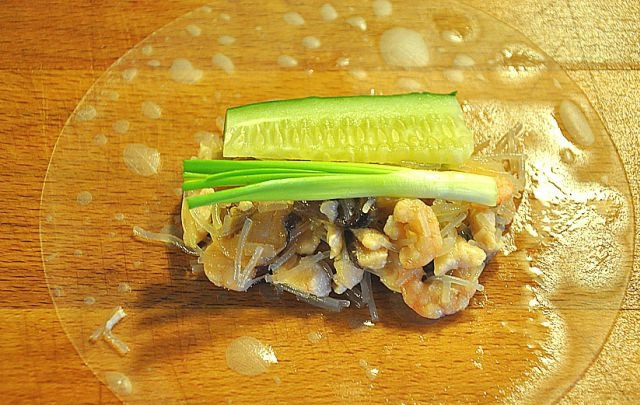 1. Этап. Рисовые роллы: 3. Отвариваем фунчозу, откидываем ее на дуршлаг, а затем перекладываем в сковороду к креветкам, курице и грибам, перемешиваем. Огурец и зеленый лук нарезаем тонкими полосками. Лист рисовой бумаги смазываем водой, затем выкладываем немного содержимого сковороды, огурец и лук.