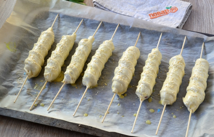 1. Этап. Сосиски в тесте на шпажках: Выложите сосиски в тесте на противень, смажьте взбитым яйцом и присыпьте кунжутными семечками.