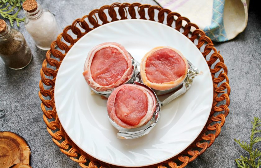 1. Этап. Свиные медальоны в беконе запеченные с овощами: Свиную вырезку зачистите от пленок, сполосните и обсушите бумажным полотенцем. Порежьте вырезку поперек на куски примерно пять сантиметров. Каждый кусок обверните беконом и закрепите полоской фольги. Каждый медальон посолите и поперчите. Дайте постоять 15 минут.