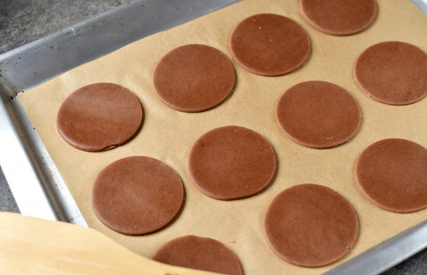 1. Этап. Печенье Новогодние олени: Раскатайте тесто между двумя листами пекарской бумаги толщиной 3-4 миллиметра. Вырубкой для печенья сделайте круглые заготовки, переложите их на противень. Если планируете украшать оленями елочку, сразу сделайте круглые отверстия вверху печенья для нитки или ленты. Отправьте противень в холодильник на 10 минут, за это время разогрейте духовку. Выпекайте печенье ровно 10 минут в режиме 