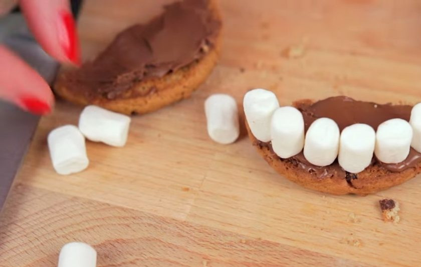 1. Этап. Снеки к Хэллоуину: 1. Для начала приготовьте челюсти из печенья. Для этого печенье разрежьте пополам, смажьте обе части нутеллой. На одну часть посредине выложите в ряд маршмеллоу в форме зубок, после чего накройте такие зубы другой половинкой печенья.