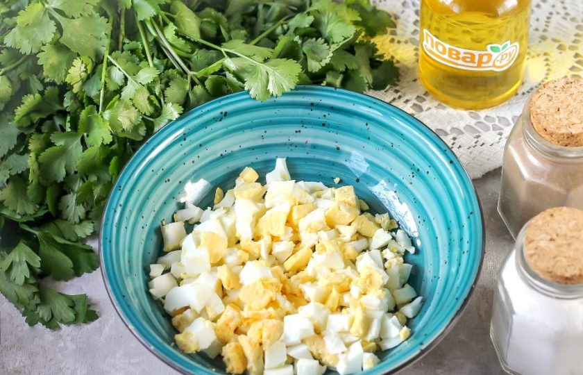 1. Этап. Салат с кальмарами и кинзой: Очистите отварные куриные яйца и промойте их в воде. Нарежьте средними кубиками и высыпьте нарезку в салатник или глубокую емкость.