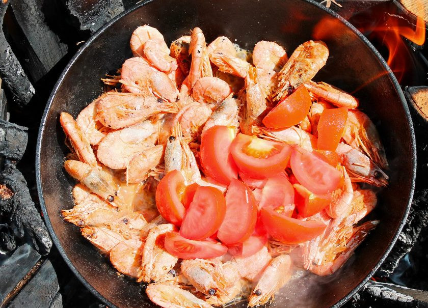 1. Этап. Креветки в казане на костре: Очищенный чеснок разрежьте на несколько частей, помидоры на дольки и выложите в казан. Готовьте еще в течение 4-5 минут, перемешивая.