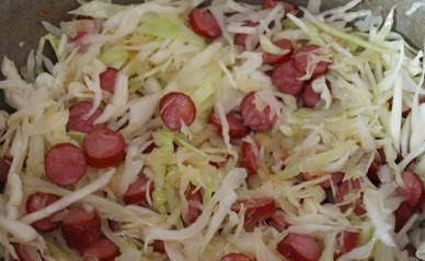 1. Этап. Тушеная капуста с колбасками: Отправляем капусту к луку с колбасками. Перемешиваем, добавляем пару ложек воды. Уменьшаем огонь, накрываем сковороду крышкой и тушим капусту минут 15.