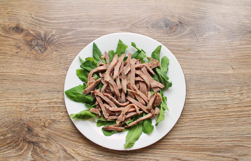 1. Этап. Салат с рукколой и языком: Рукколу выложите на дно салатника. Отварной язык порежьте мелкой соломкой и выложите ровным слоем на рукколу.