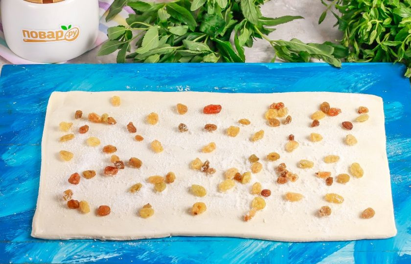 1. Этап. Слоеные палочки с изюмом и сахаром: Слоеное тесто разморозьте, если приобретали его замороженным. Раскатайте на рабочей поверхности или на доске в широкий пласт. Присыпьте сахаром и выложите изюм. Если любите пропаренные сухофрукты, то замочите изюм в кипятке за 20 минут до начала приготовления выпечки.