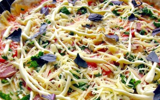 1. Этап. Пицца с копчёной колбаской: Засыпьте пиццу натёртым сыром, а сверху уложите кусочки промытого базилика.