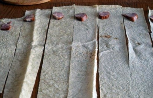 1. Этап. Треугольники из лаваша с сыром: Расстилаем лаваш, нарезаем его на полоски толщиной около 10 см. Остался маленький кусочек колбасы - вот и пустим его в дело. Разложим по кусочку колбасы на каждую полоску лаваша.