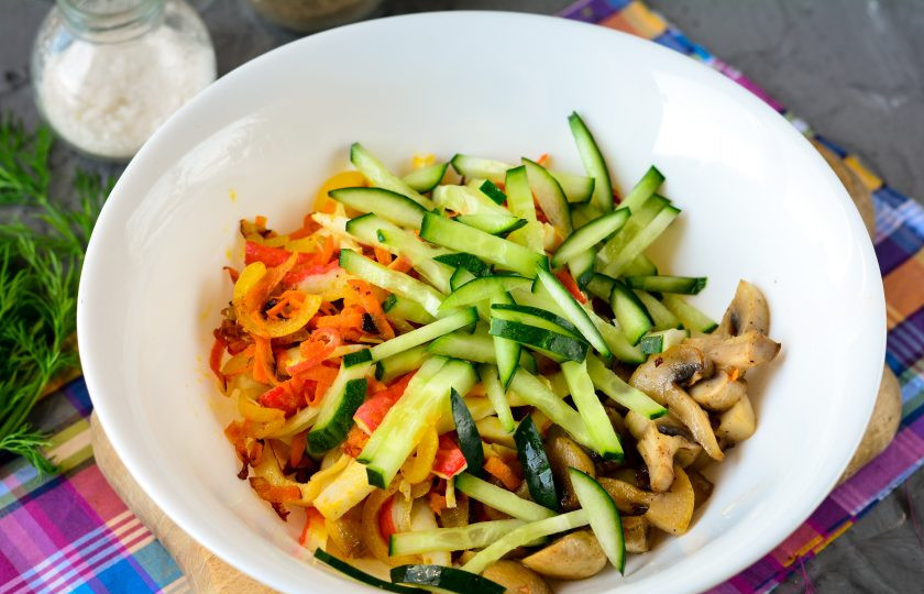 1. Этап. Салат с жареными крабовыми палочками: В пиалу выложите жареные грибы, лук с морковкой и крабовыми палочками. Также добавьте огурцы, нарезанные полосками.