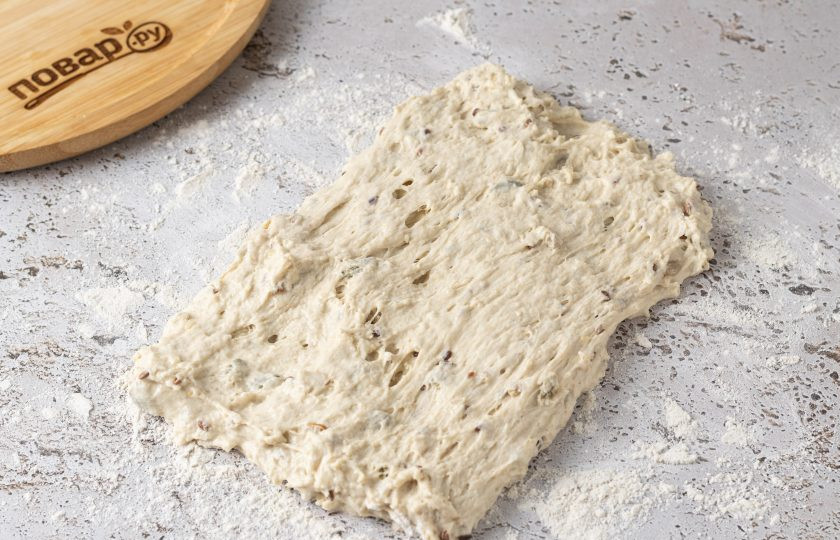 1. Этап. Ржано-пшеничный хлеб с семечками: Аккуратно с помощью силиконовой лопаточки выложите тесто на присыпанную мукой поверхность. Растяните его в прямоугольник.