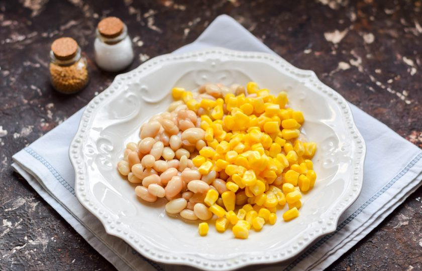 1. Этап. Салат с кукурузой, колбасой и фасолью: Консервированную фасоль промойте и переложите в тарелку. Откройте кукурузу, сок слейте, добавьте кукурузу к фасоли.