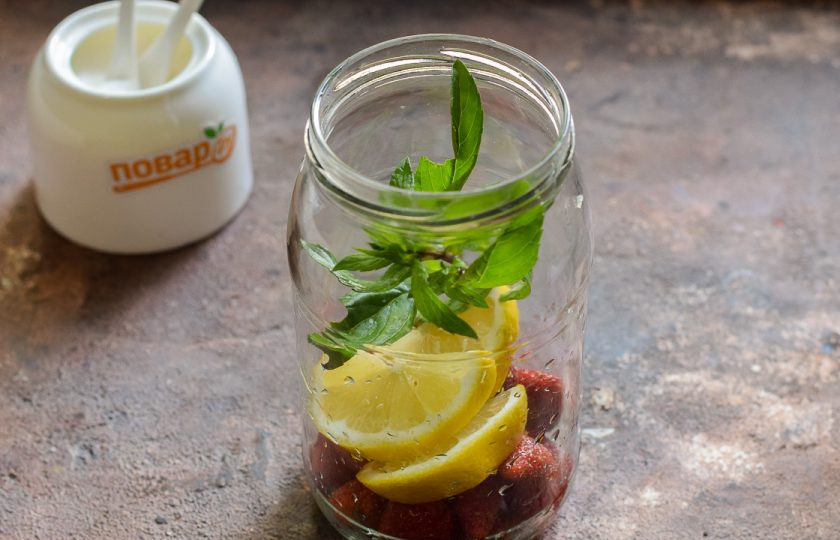 1. Этап. Компот из клубники Мохито на зиму: Банку для заготовки тщательно вымойте с пищевой содой, после ополосните хорошо под теплой проточной водой. Стерилизуйте банку и крышку удобным способом – над паром, в духовке или микроволновке. Переложите в банку клубнику, лимон и мяту.