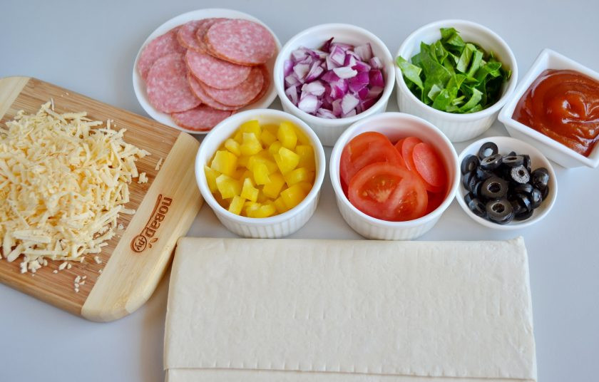 1. Этап. Закуска Пицца в стаканчиках: Разморозьте слоеное тесто при комнатной температуре. Колбасу и помидоры порежьте кружочками, лук и сладкий перец кубиками. Шпинат полосочками, оливки колечками. Сыр на крупной терке.