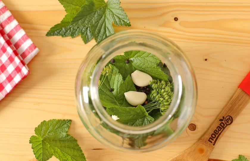 1. Этап. Огурцы с зелеными помидорами на зиму: Банки промойте и простерилизуйте любым удобным способом. Крышки прокипятите. На дно банки выложите всю зелень, очищенные зубчики чеснока, гвоздику и перец.