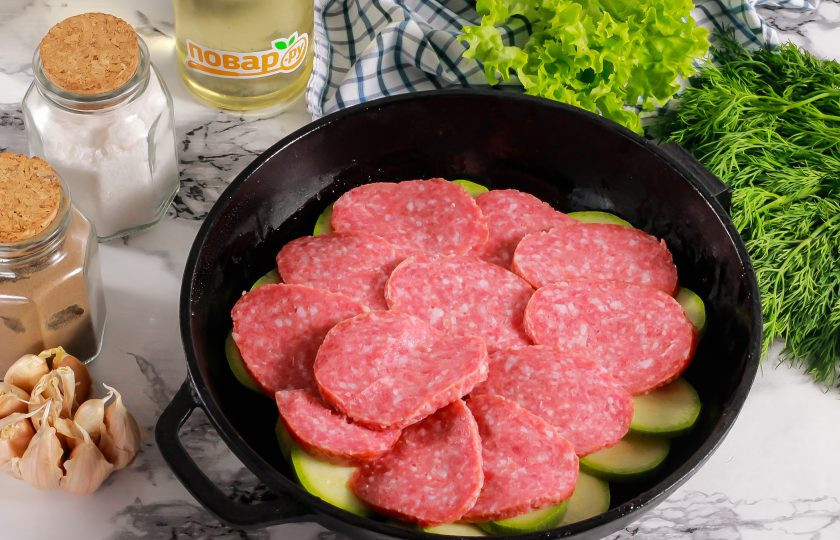 1. Этап. Кабачки запеченные с соусом и салями: Смажьте дно формы для запекания растительным маслом, выложите в нее кабачковую нарезку. Соль уже не добавляйте, так как вы ее уже солили. Нарежьте колбасу кружочками и выложите в форму.