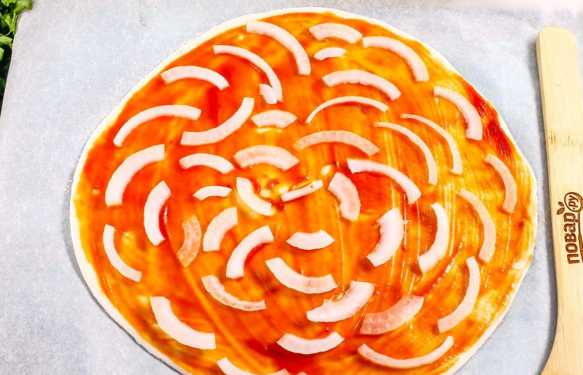 1. Этап. Пицца с колбасой из слоеного теста: Духовку разогрейте до 220 градусов. Тесто смажьте кетчупом, можно и майонезом. Половинку луковицы нарежьте тонкими ломтиками и выложите на кетчуп.