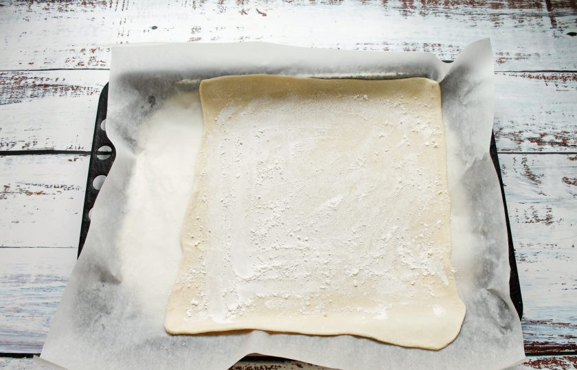 1. Этап. Слоеный пирог с абрикосами: Рабочий стол посыпьте мукой и немного раскатайте тесто в одном направлении и переложите на противень застеленный пергаментом.