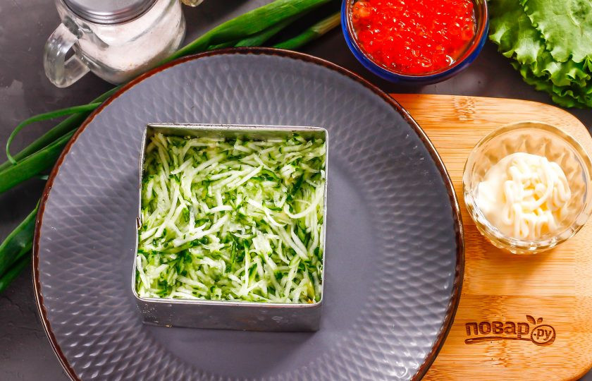 1. Этап. Слоеный салат с красной икрой: Промойте свежий огурец в воде, срежьте хвостики с овоща и натрите на терке в отдельную емкость. Посолите и оставьте нарезку на 5 минут — она выпустит жидкость. Отожмите огуречную массу от жидкости и выложите в кольцо или квадрат.