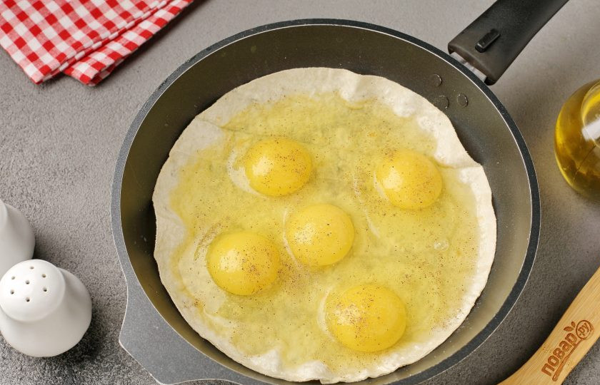 1. Этап. Яичница с помидором и сыром в лаваше на сковороде: Разогрейте сковороду и смажьте маслом. Выложите круглый лаваш. Если у вас нет лаваша подходящей формы, то при помощи кухонных ножниц, просто вырежьте заготовки из обычного квадратного. Сверху на лаваш разбейте необходимое количество яиц. Посолите и поперчите по вкусу.