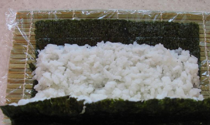 1. Этап. Роллы с икрой летучей рыбы: Застелите бамбуковый коврик пищевой пленкой. Выложите на коврик один лист нори. Мокрыми руками равномерно распределите рис (1 см. до края оставьте свободным). Ловким движением переверните лист нори, чтобы рис оказался снизу.