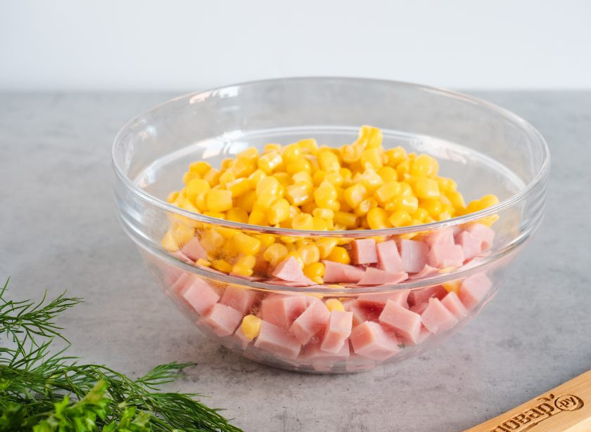 1. Этап. Салат с ветчиной, яйцом и кукурузой: Ветчину нарежьте небольшими кубиками и положите в салатник. С кукурузы слейте рассол и добавьте к ветчине.