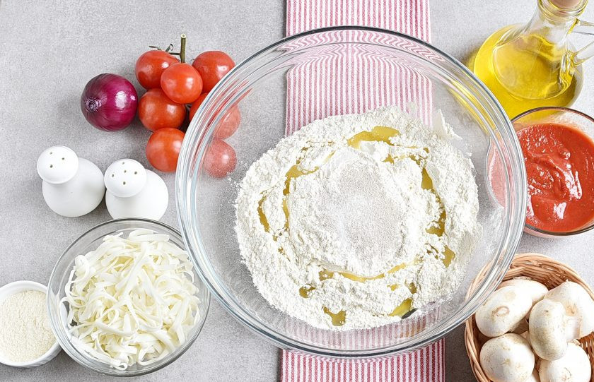 1. Этап. Пицца с шампиньонами: Смешайте просеянную муку с солью и сухими дрожжами. Влейте две столовых ложки оливкового масла. Постепенно подливая теплую воду, замесите плотное, но нежное тесто.