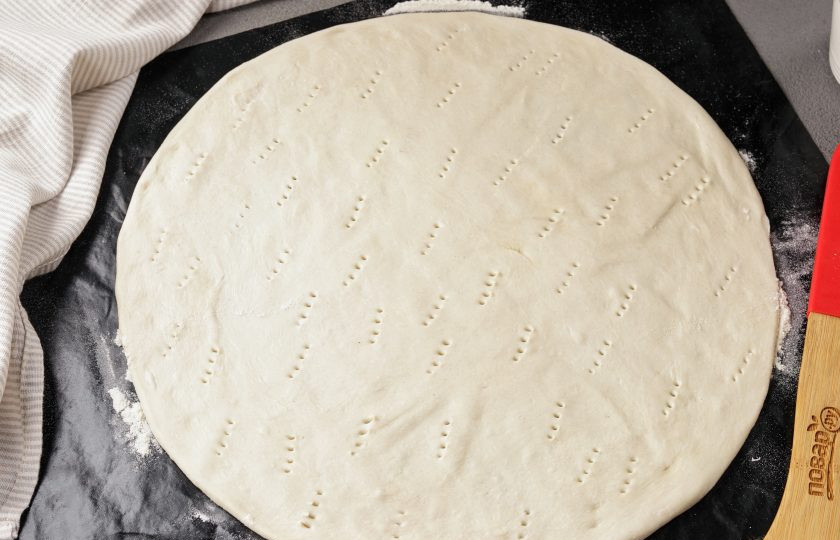 1. Этап. Сладкая пицца с ананасами: Пергамент или специальный кулинарный коврик присыпьте мукой. Выложите тесто и тонко раскатайте. Наколите тесто вилкой.