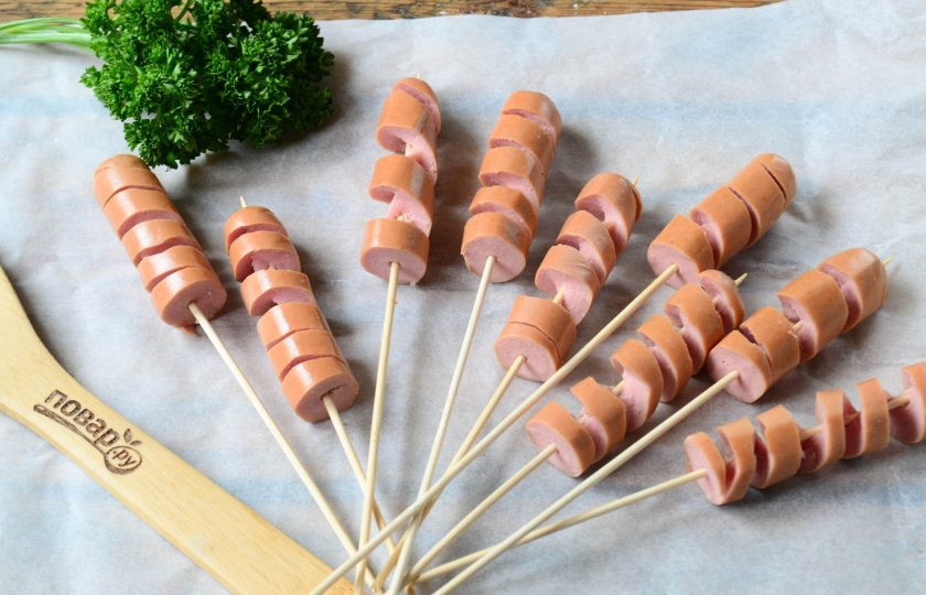 1. Этап. Сосиски в тесте на палочке Твистер: Каждую сосиску разрежьте пополам и оденьте на деревянную шпажку. Затем острым ножом сделайте надрезы по спирали, доходя до шпажки. У вас получится спираль из сосиски, максимально растяните ее.