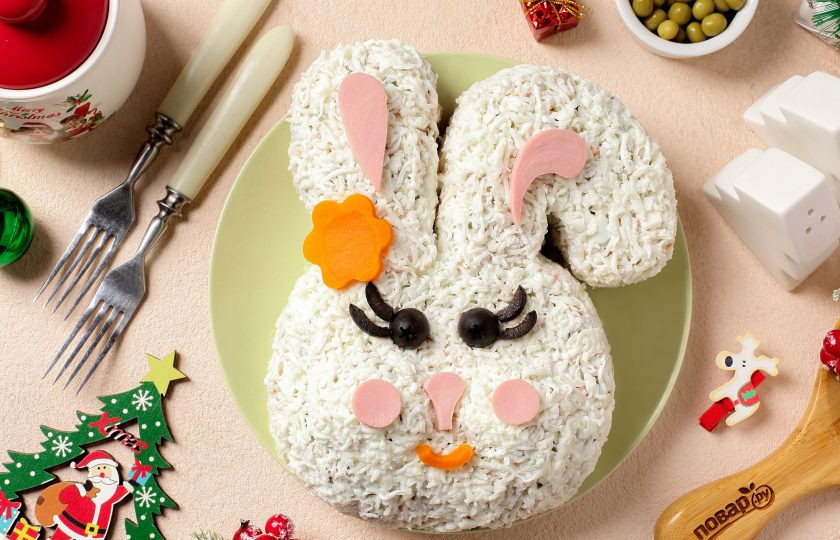 1. Этап. Салат Оливье на год Кролика: Белки варёных яиц натрите на мелкой тёрке и выложите сверху, а дальше просто включаем фантазию и делаем мордочку нашему забавному зверьку. Ушки, носик и щёчки я сделала из варёной колбасы. Глаза из маслин, а ротик и бантик на ушке из варёной морковки. Салат 