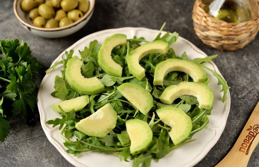 1. Этап. Салат с курицей, авокадо и рукколой: Авокадо почистите от косточки и кожуры, нарежьте тонкими ломтиками. Подготовьте плоскую тарелку, выложите на нее рукколу, следующим слоем выложите авокадо.