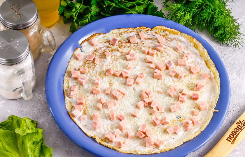 1. Этап. Блинный торт с ветчиной и сыром: Выложите блин на тарелку и смажьте его сметаной любой жирности либо майонезом. Нарежьте ветчину мелкими кубиками, а сыр натрите на терке с мелкими ячейками. Выложите на сметану часть нарезки ветчины.