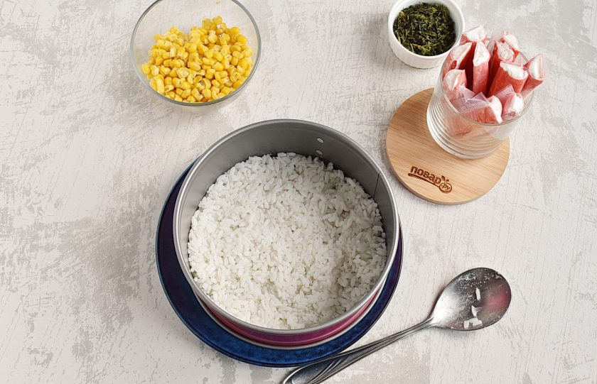 1. Этап. Крабовый салат с рисом и кукурузой: На блюдо установите разъемное кольцо и выложите отварной рис первым слоем, хорошо утрамбовывая.