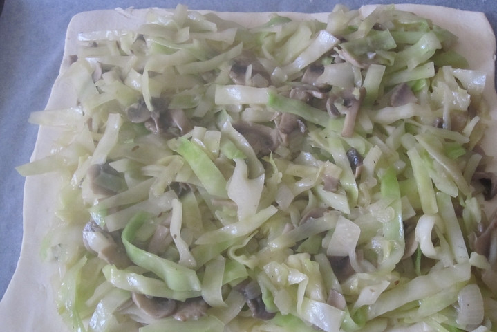 1. Этап. Слоеное бездрожжевое тесто с капустой: Тонко раскатайте пласт слоеного теста. Выложите его на противень, застеленный бумагой для выпечки. Разложите на тесто начинку.