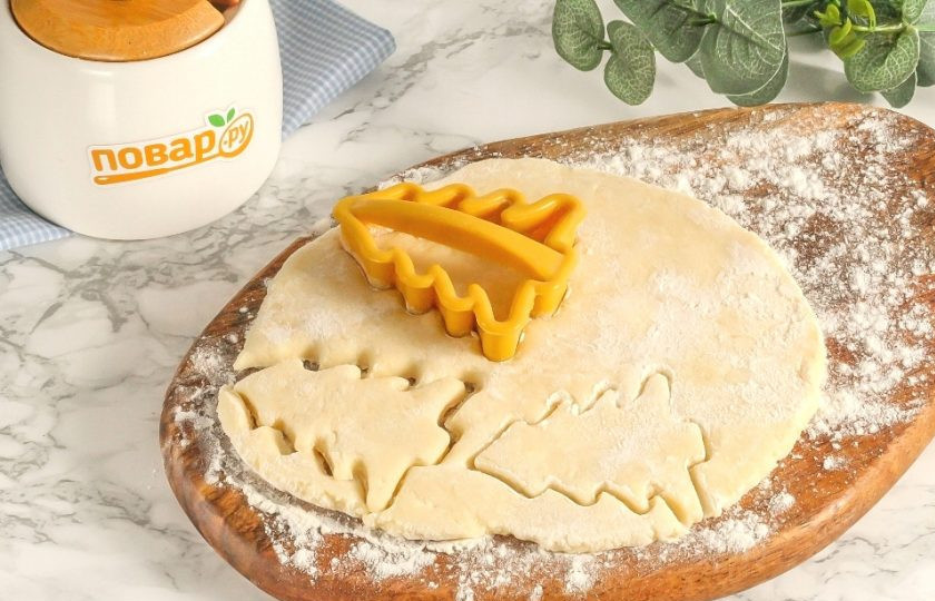 1. Этап. Творожное печенье «Елочки»: Извлеките тесто и разделите его на три части. Раскатайте каждую из них в небольшой круглый пласт шириной не менее 1 см. Меньше не раскатывайте, иначе не сможете отделить вырубленную заготовку от рабочей поверхности, так как тесто слишком мягкое и податливое. Обязательно подпыляйте доску мукой! С помощью вырубки вырежьте из теста елочки.