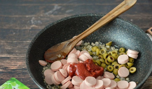 1. Этап. Горячая закуска из сосисок Солянка: Сосиски освободите от оболочки, нарежьте кружочками. Также кружочками нарежьте оливки. В сковороду к луку с огурцами добавьте сосиски, оливки, томатную пасту.