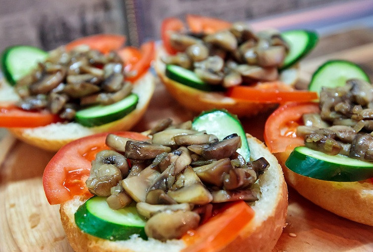 1. Этап. Бутерброды с шампиньонами и сыром: Теперь кладем в каждую выемку по несколько ломтиков помидора и огурца, сверху на овощи выкладываем обжаренные грибы с луком.