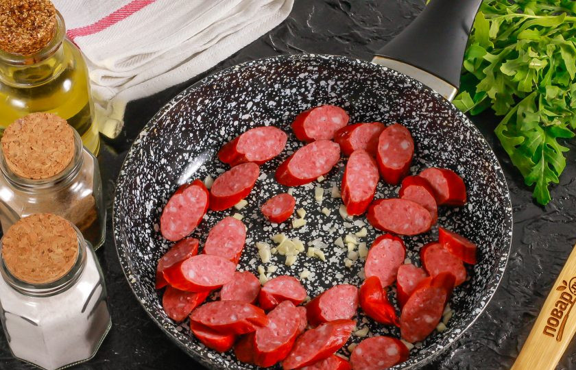 1. Этап. Макароны с охотничьими колбасками: Чесночные зубчики очистите от шелухи и промойте в воде. Нарежьте мелко или спрессуйте через специальный девайс. Добавьте чесночную массу в сковороду, влив растительное масло. Поместите емкость на плиту и прогрейте. Нарежьте охотничьи колбаски ломтиками и добавьте в сковороду. Обжарьте примерно 2 минуты.