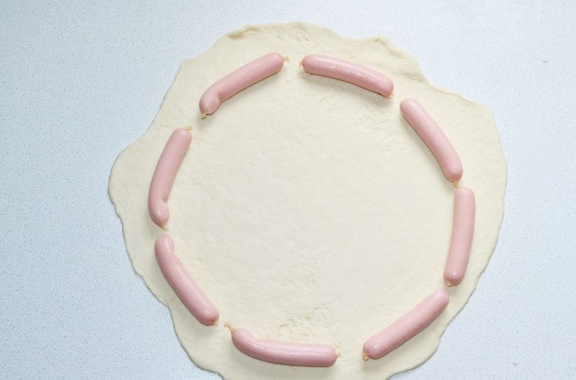 1. Этап. Пицца с сосисками по краям: Тесто разделите на 2-3 части. Раскатайте тонким пластом и выложите по краям сосиски.