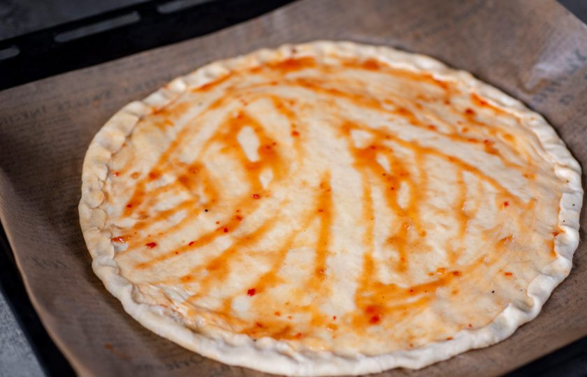 1. Этап. Мексиканская острая пицца: Противень застелите пергаментом для выпекания, и на нем растяните тесто руками, придавая ему форму круга — это основа для будущей пиццы. Поверхность основы смажьте тонким слоем соуса чили.