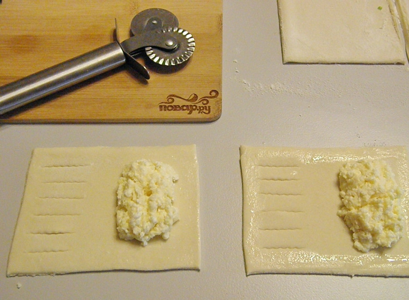 1. Этап. Слойки с творогом из готового слоеного теста: Порежьте тесто, не раскатывая на прямоугольники. С одной стороны сделайте красивые надрезы. Весь периметр смажьте белком, чтобы лучше склеился край слойки. В центр выложите по столовой ложке творога.