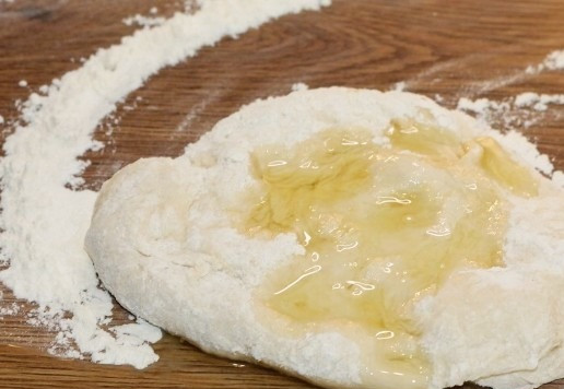 1. Этап. Морские узелки с чесночным маслом: 6. Остальную муку высыпьте на столешницу. Выложите тесто из миски и продолжайте замес. Затем полейте тесто растительным маслом (2-3 ст. л.). Вмешивайте масло в тесто. Соберите тесто в ком и округлите. Тесто должно получится мягкое и не липнущее к рукам.