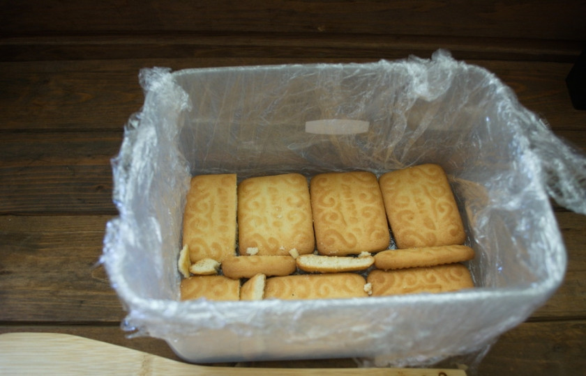 1. Этап. Клубнично-творожный торт без выпечки: Емкость застелите пищевой пленкой. На дно уложите в один ряд печенье. Пустые места закройте кусочками печенья.