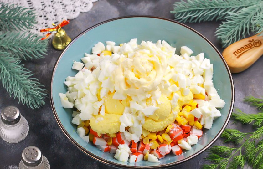 1. Этап. Крабовый салат на Новый год 2025: Отделите белки двух яиц в отдельную пиалу — они вам потребуются для украшения салата. Остальные яйца нарежьте таким же кубиком и отправьте к остальным ингредиентам в емкость. Посолите.